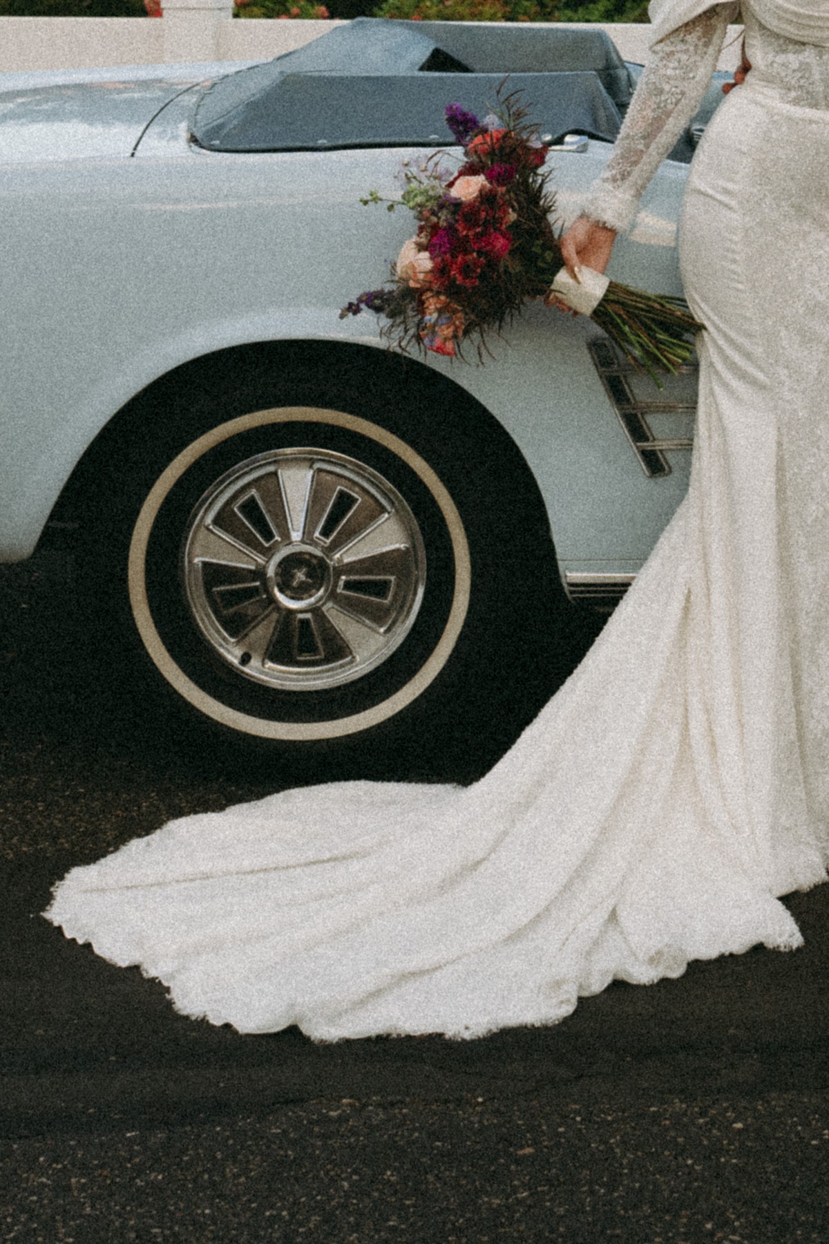 A bride’s bouquet and dress beside a vintage convertible